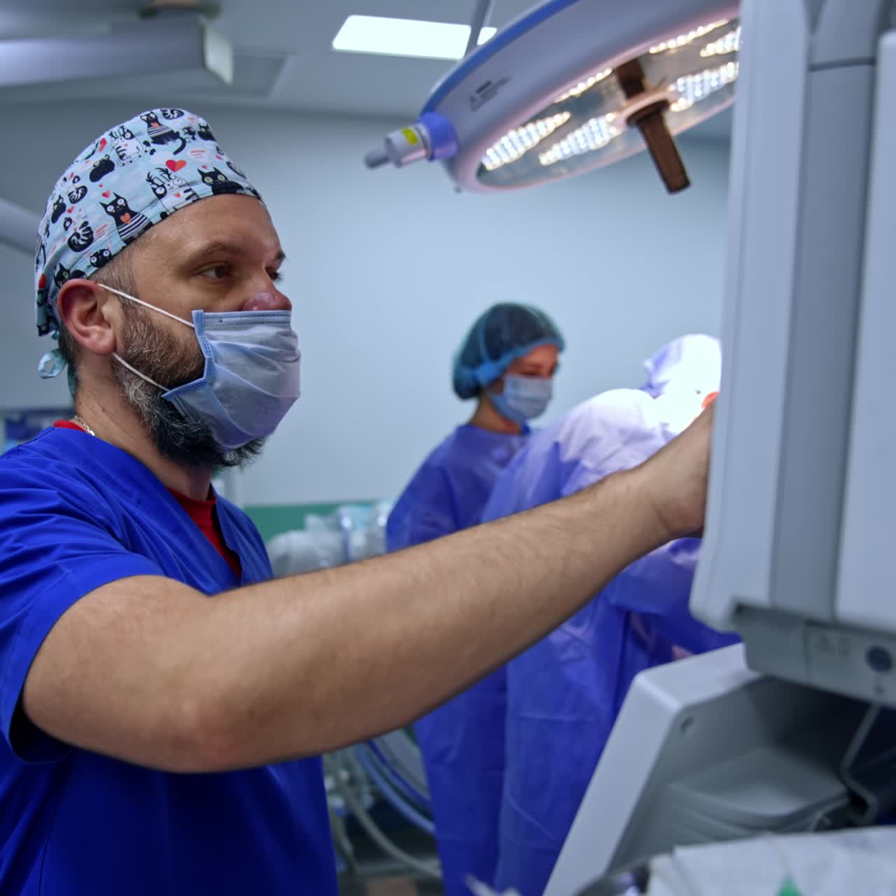 Caucasian male anesthesiologist stands near the lung ventilator machine controlling the patient's condition. Surgeons operate at backdrop
