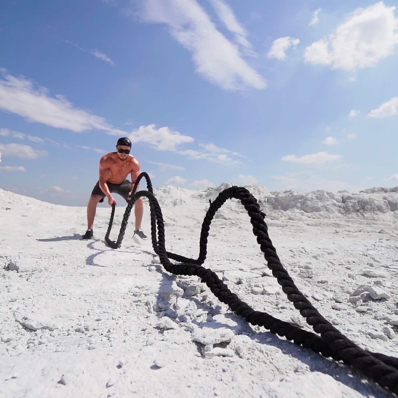 Male athlete exercising outdoors around white hills at daytime. Active physical workout. Strong man in sunglasses has a workout with a rope on the ground.