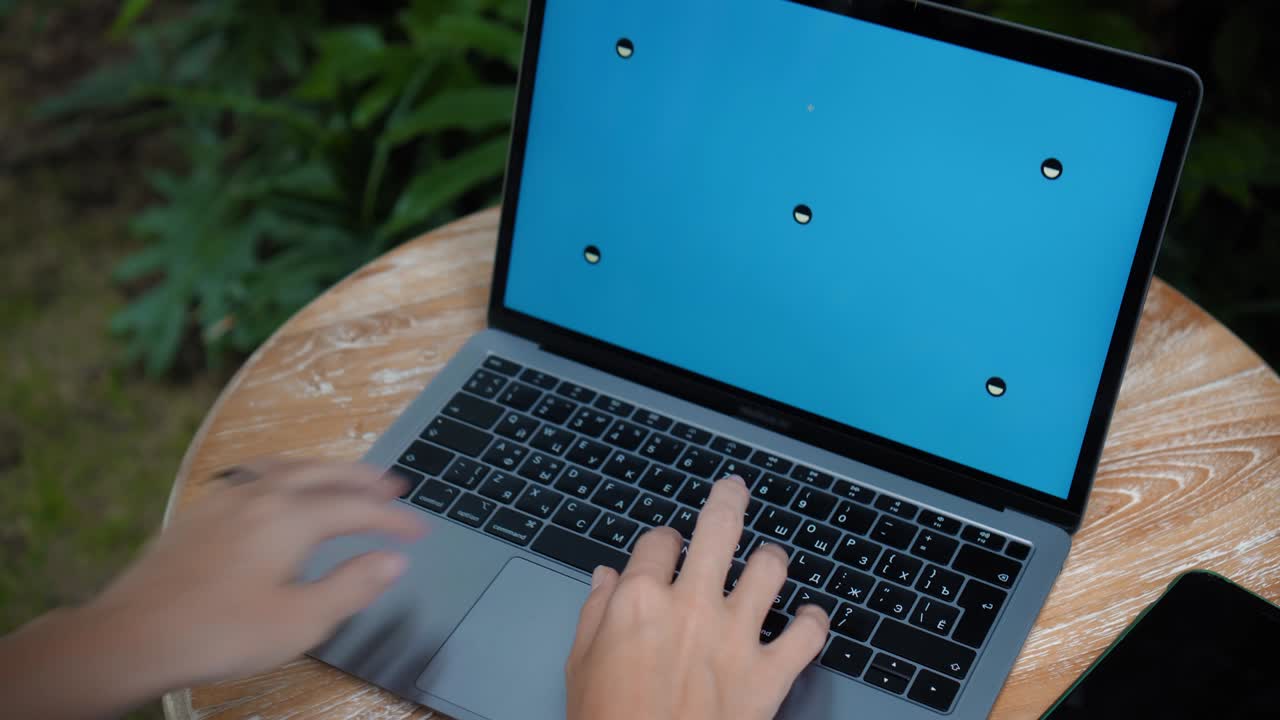 un joven europeo en un vestido azul remotamente en línea trabajando en una computadora portátil con pantalla azul en el patio trasero con plantas verdes