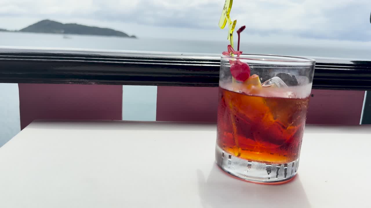 A hand places a Negroni cocktail on a table overlooking a serene beach view in Phuket, Thailand