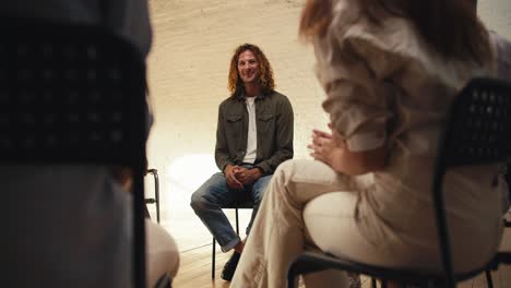 a man with curly red hair in a green gray sweater expresses his problem at groove therapy in a white brick hall. other participants in a group therapy meeting applaud the man