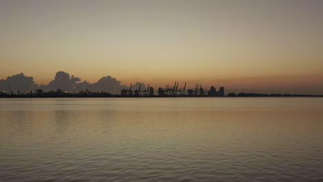 Vista-Aérea-Moviéndose-Hacia-Grúas-Portuarias-De-Envío-Con-Marcador-De-Marea-De-Silueta-En-La-Costa-De-Miami-Al-Atardecer