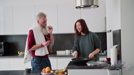 Happy-elderly-man-with-gray-hair-and-a-lush-beard-brushes-his-teeth-in-the-kitchen-while-his-adult-daughter-prepares-breakfast.-Happy-brunette-girl-in-a-green-sweater-dances-with-her-elderly-father-with-gray-hair-while-preparing-breakfast-in-the-kitchen-in-a-modern-apartment