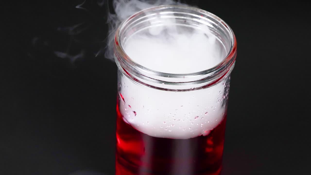 una secuencia que muestra hielo seco reaccionando en un líquido rojo, creando niebla y burbujas en un vaso de vidrio