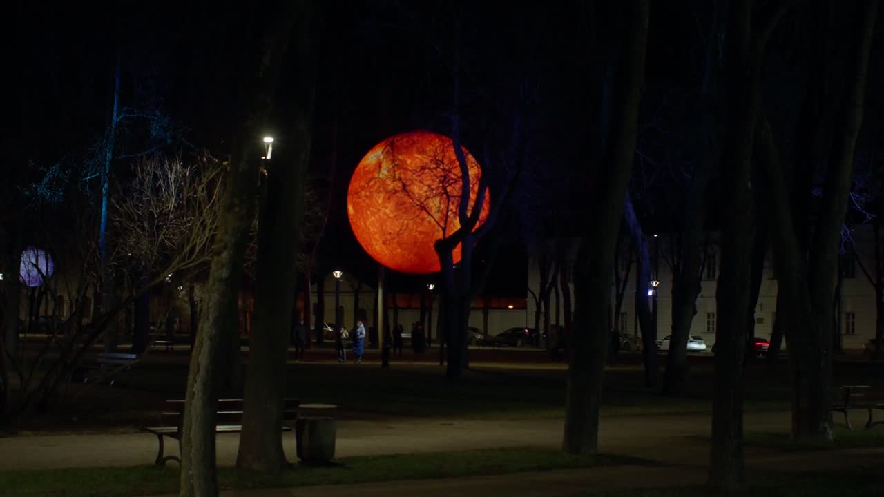 Esfera naranja brillante instalada en un parque oscuro durante un festival de invierno. La silueta de árboles desnudos enmarca una obra de arte luminosa similar a la luna. Gente caminando al fondo de la exposición nocturna.