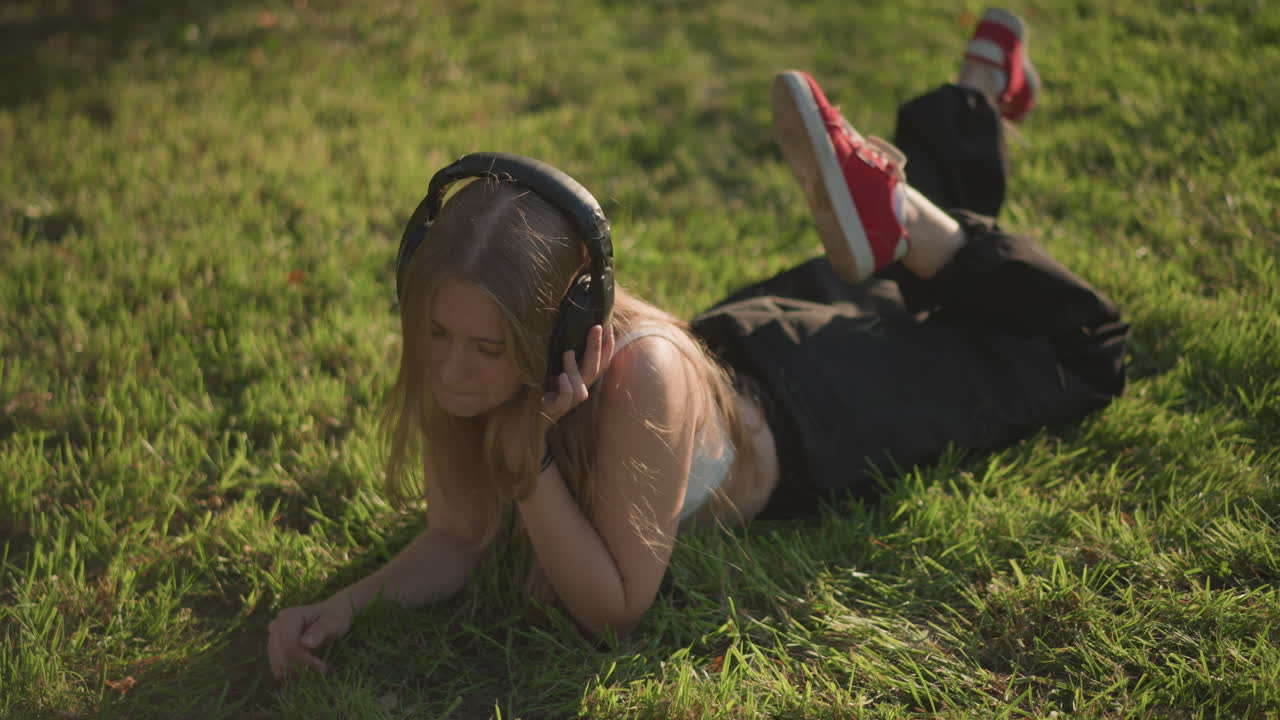 young caucasian woman lying on grass, sunlit afternoon, oversized headphones, warm golden light, casual black pants and red sneakers, gentle smile, slow head sway, peaceful leisure, serene youthful