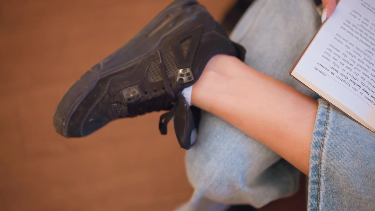 Primo piano della gamba di una giovane donna che indossa jeans blu e sneakers nere mentre legge un libro. La scena mostra il dettaglio della texture del tessuto denim, calzini bianchi ai piedi e un'illuminazione soffusa sulla tonalità della pelle.