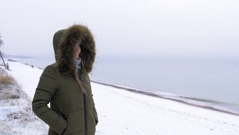 Young-woman-in-green-winter-clothes-stands-on-the-sandy-shore-of-the-Baltic-sea-beach,-grass,-medium-jib-shot-up