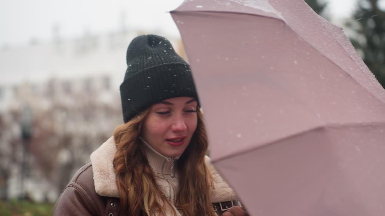 Gros plan d’une jeune fille portant un bonnet en maille noir et une veste en peau de mouton marron, tenant un parapluie tandis que le vent souffle doucement avec de légères chutes de neige, des mèches de cheveux en mouvement, un arrière-plan doux et paisible avec une ambiance hivernale