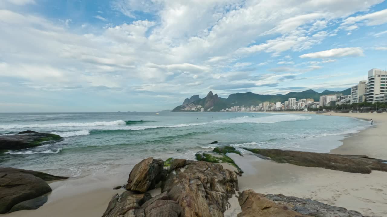 Time lapse early morning Ipanema beach in Rio de Janeiro with rocks scattered in the foreground and the Two Brothers mountain in the background against a blue cloudy sky