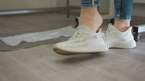 close-up view of lady in fitted jeans adjusting stylish white sneakers while standing on wooden floor indoors, with part of gray floor mat and furniture visible in soft background