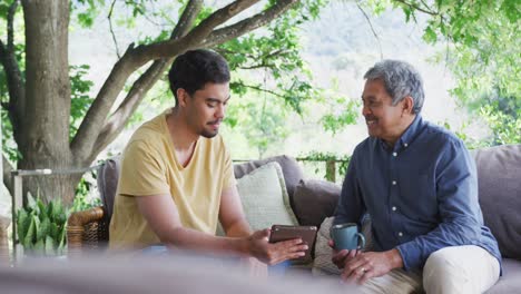 Cheerful-father-and-son-talking-while-sitting-with-tablet-pc-and-coffee-mug-on-sofa-at-patio