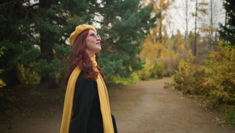 elegant girl with yellow beret and muffler smiles warmly, touching her red wavy hair as she walks through park observing her surroundings while strolling in a vibrant garden