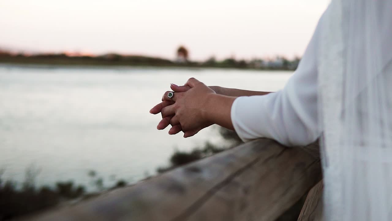 Close-up of the bride's folded hands resting on a wooden railing watching the sunset over a lake