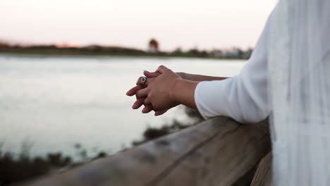 Close-up-of-the-bride's-folded-hands-resting-on-a-wooden-railing-watching-the-sunset-over-a-lake