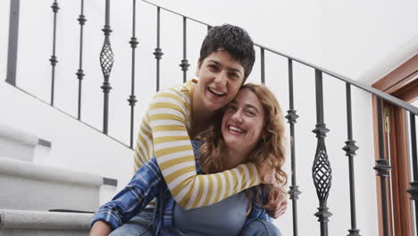 una feliz pareja de lesbianas caucásicas sentadas en las escaleras, abrazándose y sonriendo en una casa soleada