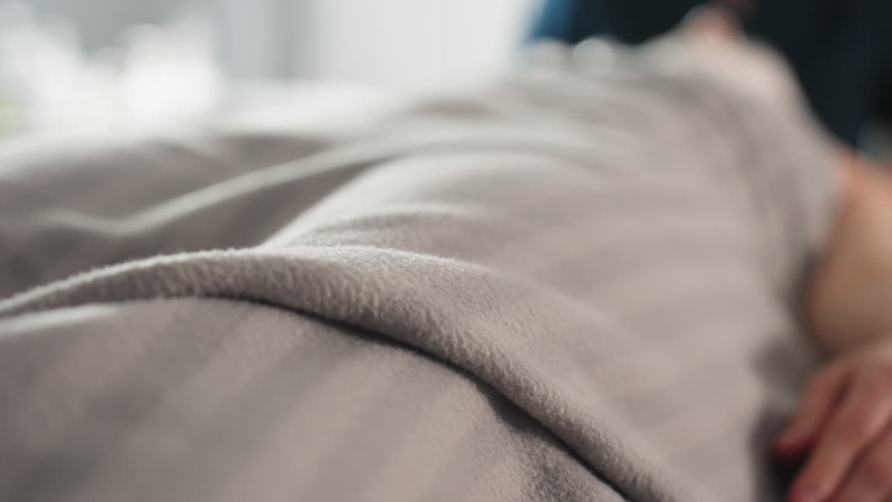 Hand of spa guest peeks from under soft blanket on massage bed while therapist works on facial in blurred background, conveying tranquil treatment scene during beauty therapy session