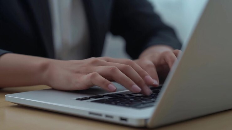 Tippende Erwachsenenhände, die Tasten auf einem silbernen Laptop am Schreibtisch drücken, arbeiten in Blazerärmeln.