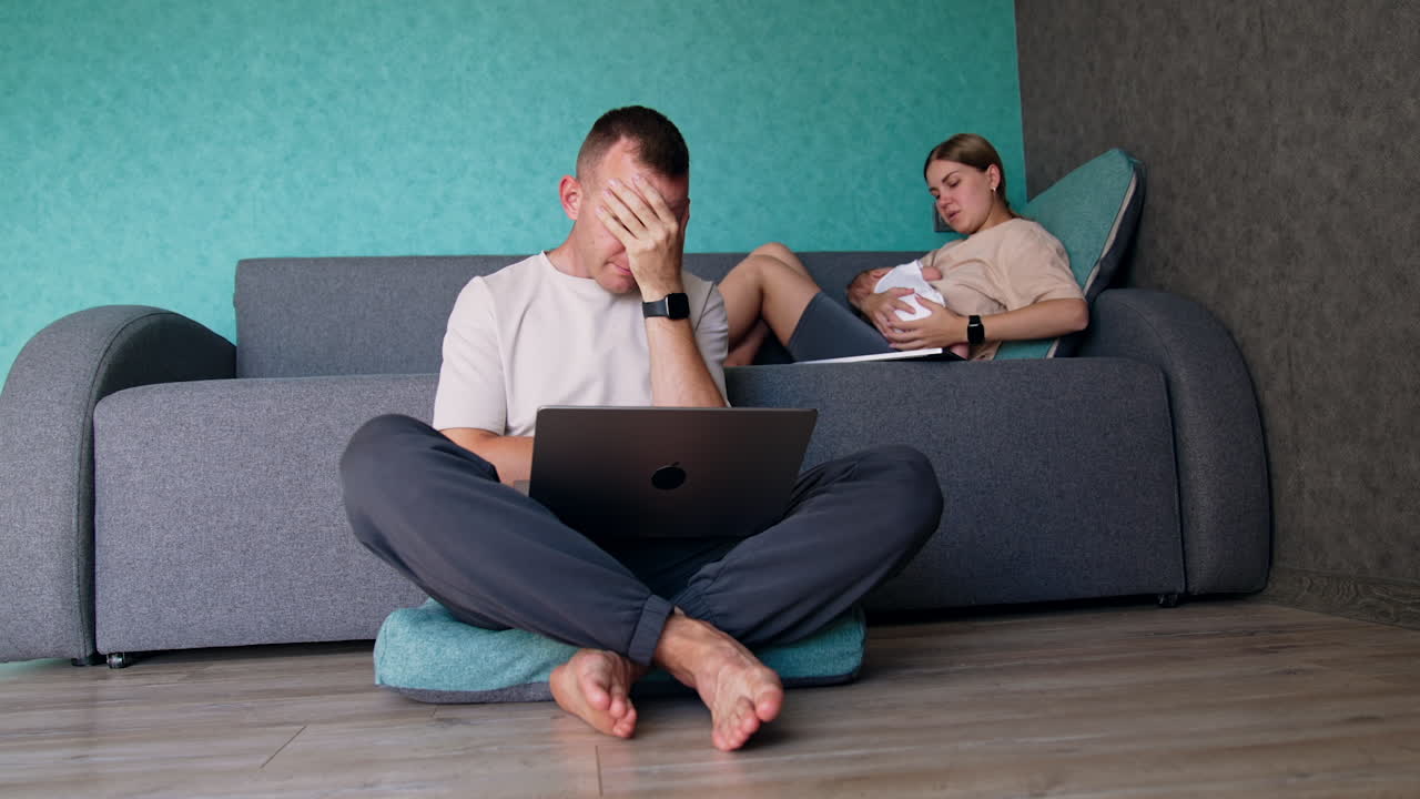 Tired man sitting near the sofa in front of his laptop. Woman sits on the sofa with child in hands. New parents working remote.