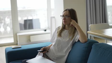 A-young-woman-sitting-on-a-sofa-with-a-TV-remote-control-in-her-hands-and-switching-channels