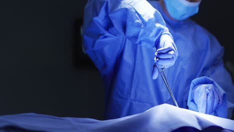 Mid-section-of-surgeon-wearing-protective-clothing-holding-scissors-in-operating-theatre