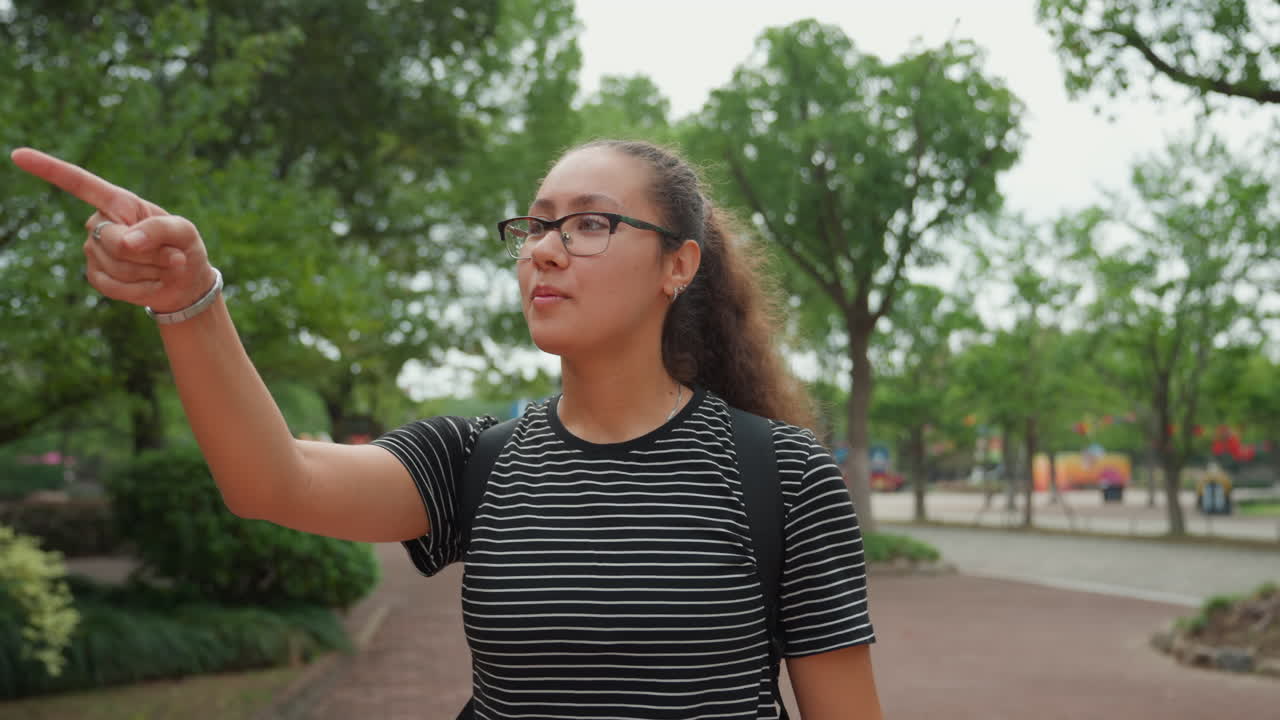 Giovane donna in un parco che esplora un sentiero e indica gli alberi, con un'aria da studentessa casual, zaino e occhiali, capelli ricci raccolti, camminando lungo un sentiero pavimentato vicino a un parco giochi, luce diurna nuvolosa.