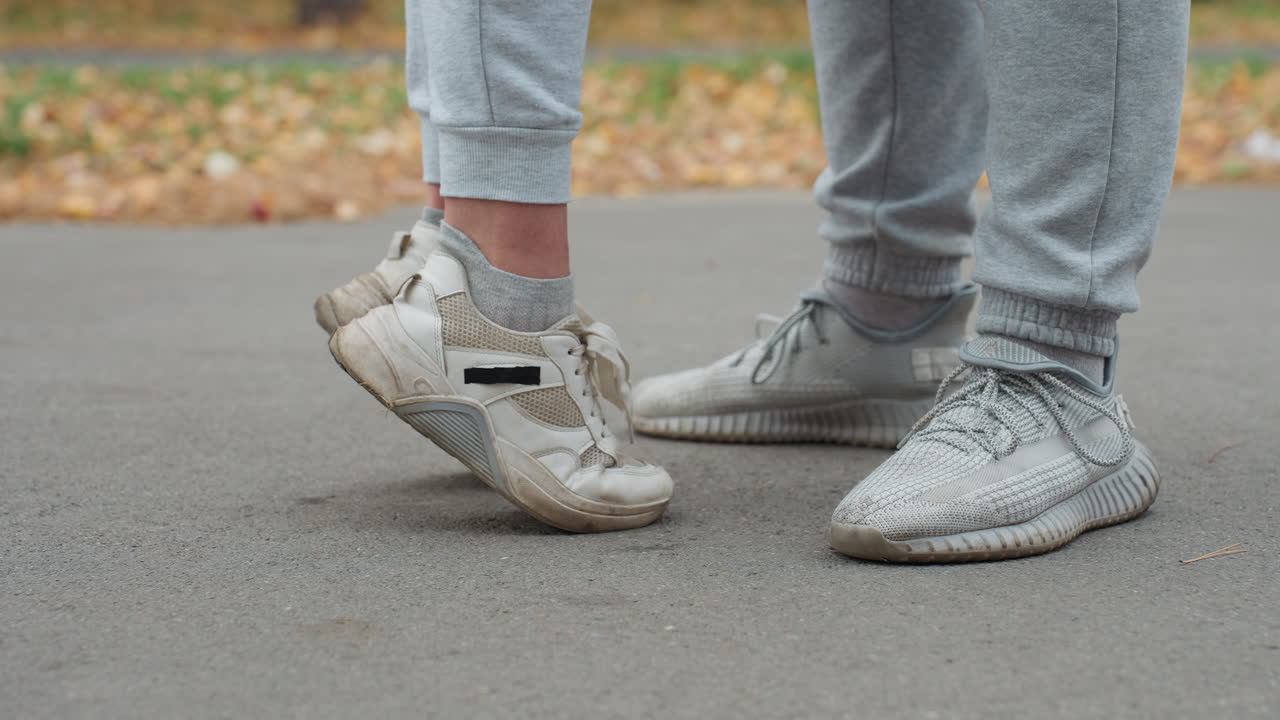 Side leg view of two people in joggers and sneakers standing on pavement as one person stretches upward lifting feet off ground, with blur background of fallen dry foliage