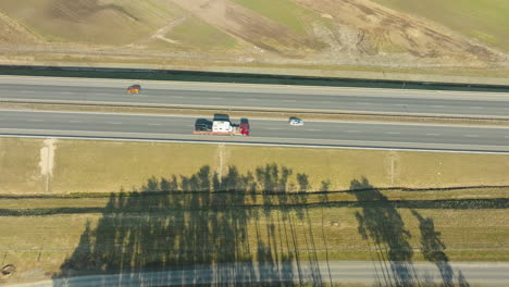 shadows stretch across a highway where colorful vehicles are spaced out, journeying along a road bordered by changing terrain