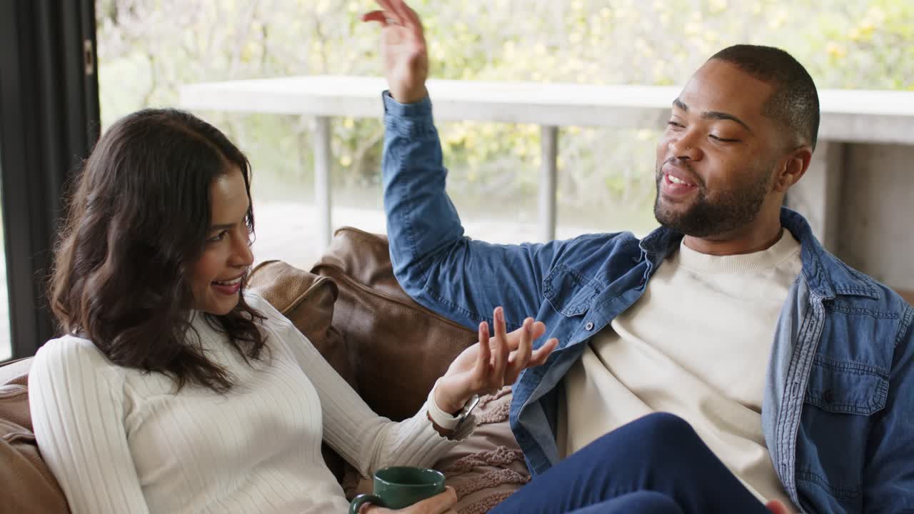 Diverse couple holding green mug, gesturing while exchanging ideas on sofa in living room