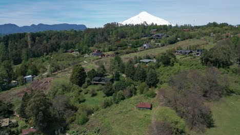 drone shot above hillside cabins and villas amid lush green forests with the snow-capped villarrica volcano in the background in chile
