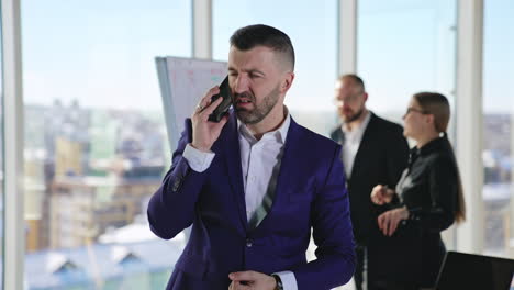 emotional businessman speaking on the smartphone. man speaks and gestures actively. employees standing at the chart in the backdrop.