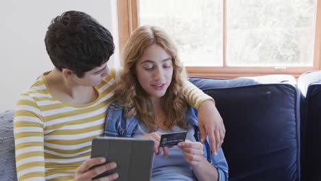 feliz pareja de lesbianas caucásicas abrazándose en el sofá, usando tableta y tarjeta de crédito en la casa soleada