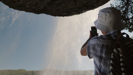 a woman standing under a waterfall steinsdalsfossen takes a photo above it hangs a rock the majestic