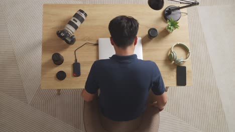 top view of asian male color grading walking into the workspace sitting down and using a laptop next to the camera editing the video at home