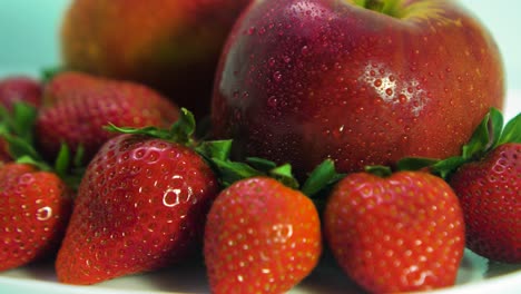 Fresh-big-red-tasty-ripe-strawberries-and-red-apples-covered-by-water-droplets-rotates-slowly-on-a-white-plate-on-light-blue-background,-healthy-food-concept,-extreme-closeup-shot,-camera-rotate-right