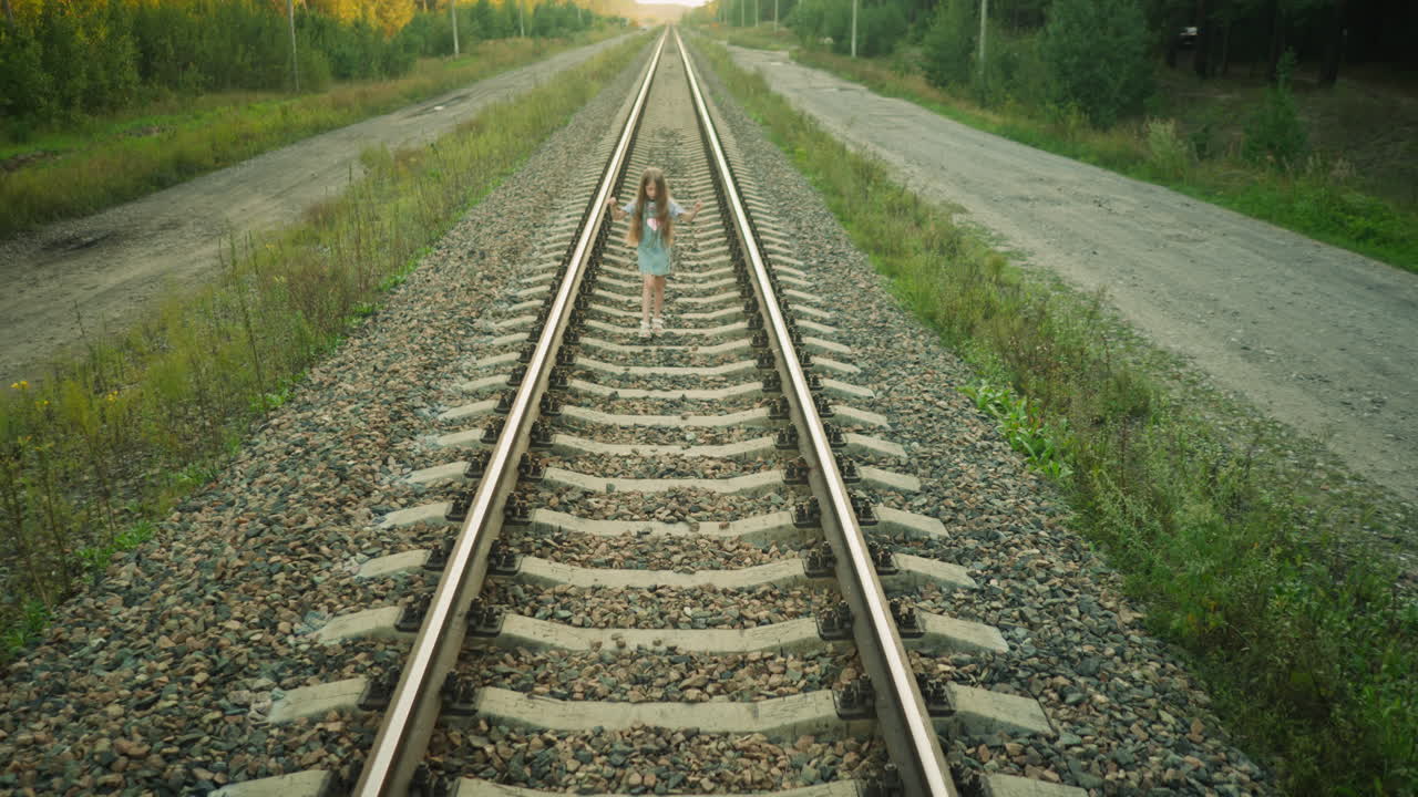 Kid walking along railway track with one hand lifted in countryside setting, dressed in denim dress and striped shirt, surrounded by gravel, wild greenery, and dirt road