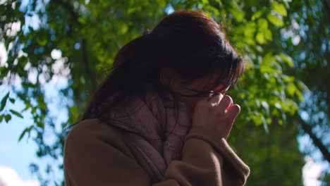 shot of woman crying, hiding face in hands while visiting a park surrounded by green trees during morning time