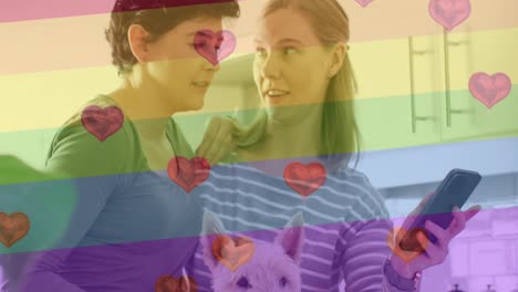 Animation-of-heart-emojis-and-rainbow-flag-over-caucasian-female-couple-using-smartphone