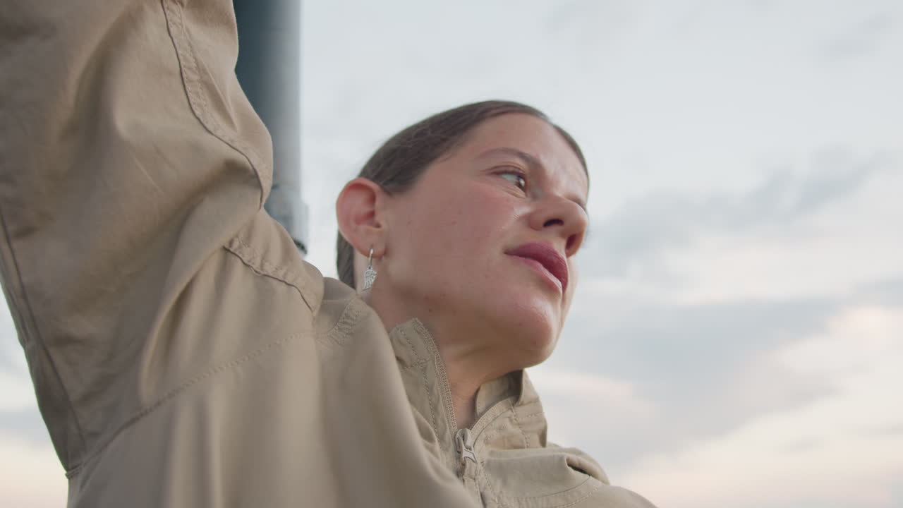 female pilot gripping sturdy support pole inside colorful hot air balloon basket at sunrise wind tousled hair framing focused gaze panoramic farmland visible below