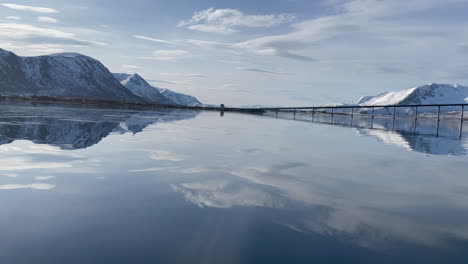 View-of-Risoyhamn-Bridge-from-the-pristine-blue-lake-in-Lofoten,-Andøya-Vesterålen,-Norway-surrounded-by-beautiful-snow-covered-mountains-on-all-sides