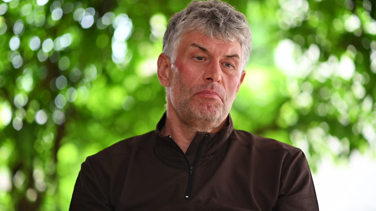 Close up of a middle aged man sitting outdoors and talking casually in natural sunlight with green foliage in the background