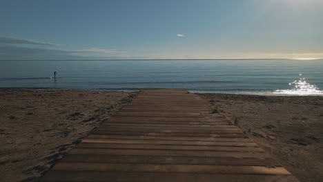 Drone-shot-of-beach-pathwalk,-moving-forward-into-the-sea-with-paddler-passing-by