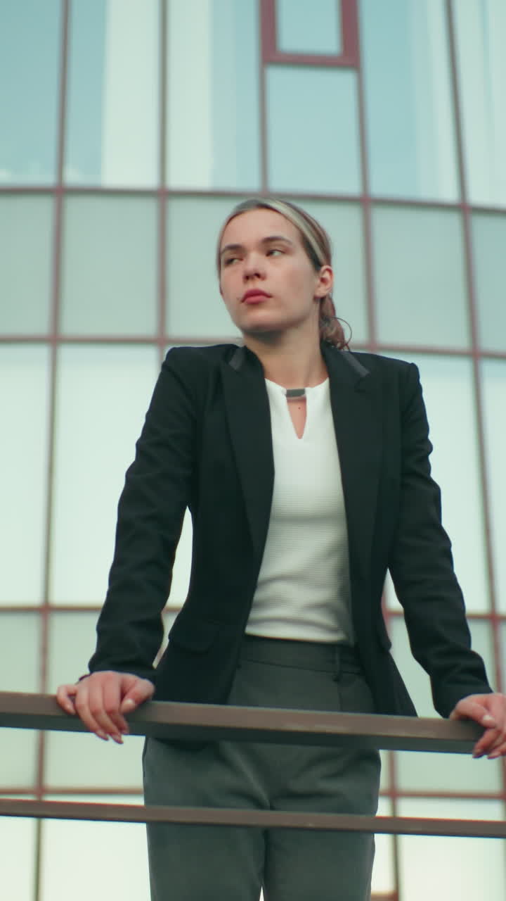 Professional girl in formal outfit leans on iron railing, gazing into distance with focused expression, beside lamp post and CCTV camera, modern glass office building in background