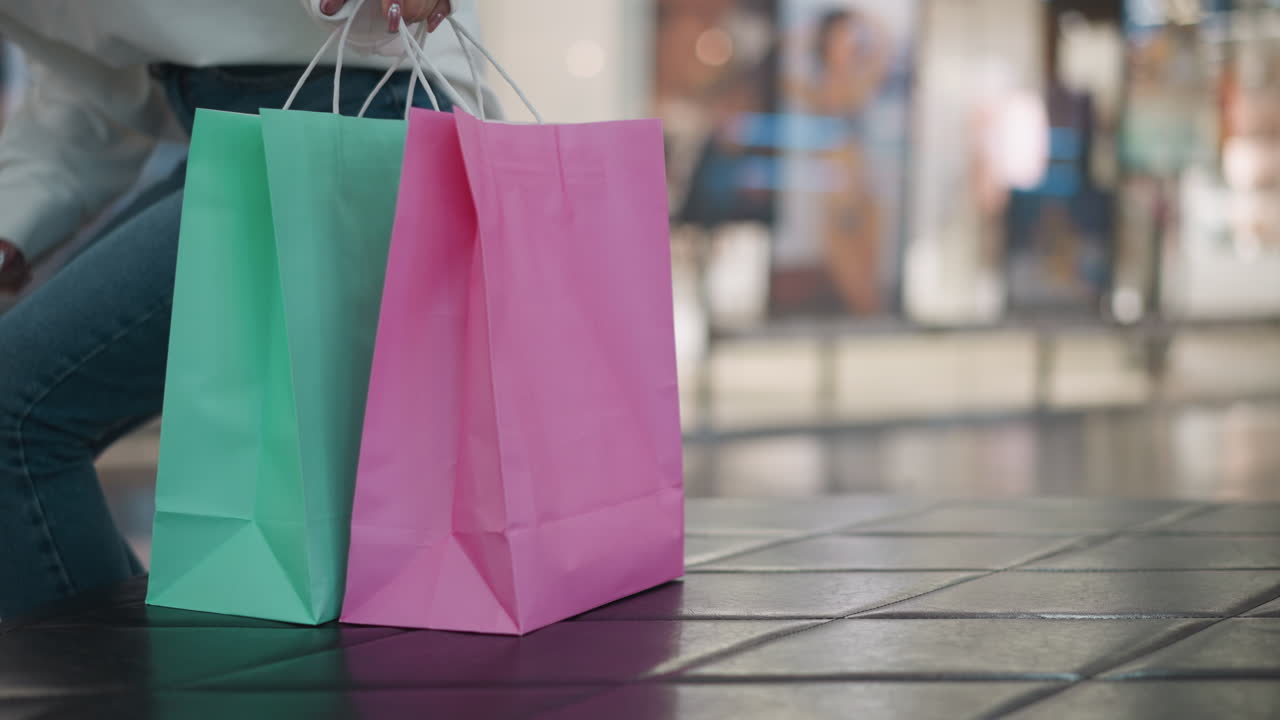 bolsas de compras de menta y rosa colocadas en una superficie de azulejos, ligeramente inclinadas, con un fondo borroso del centro comercial, una mujer en vaqueros se sienta cerca en el asiento de cuero