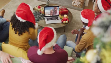 Diverse-family-with-santa-hats-using-laptop-for-christmas-video-call-with-happy-couple-on-screen