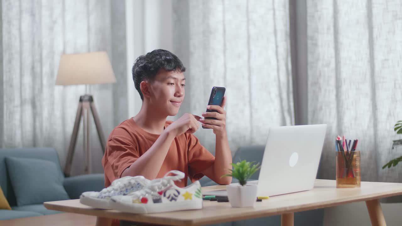 아시아 소년 신발 디자이너가 집에서 노트북으로 작업하는 동안 스마트폰을 보고 있습니다.
