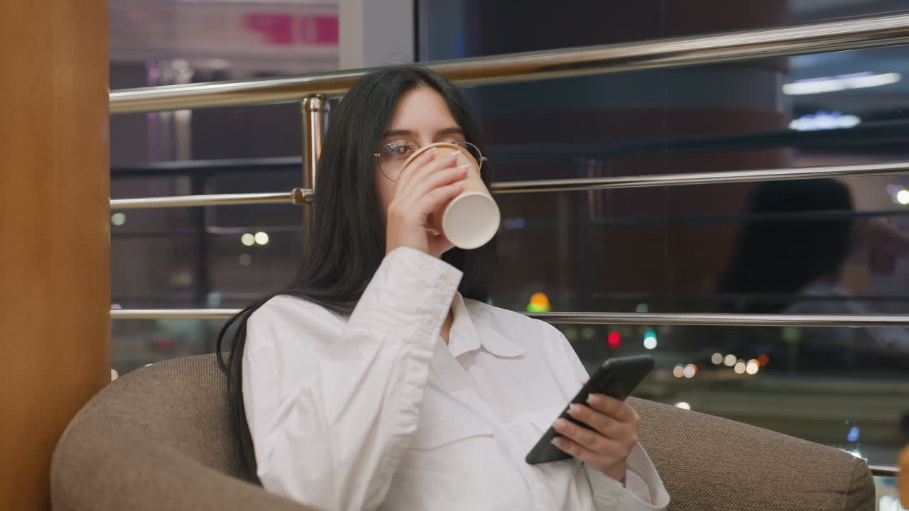 elegant lady holding coffee cup drops it gently while using smartphone seated indoors with shiny iron railing and busy night urban lights visible through large glass panel in background