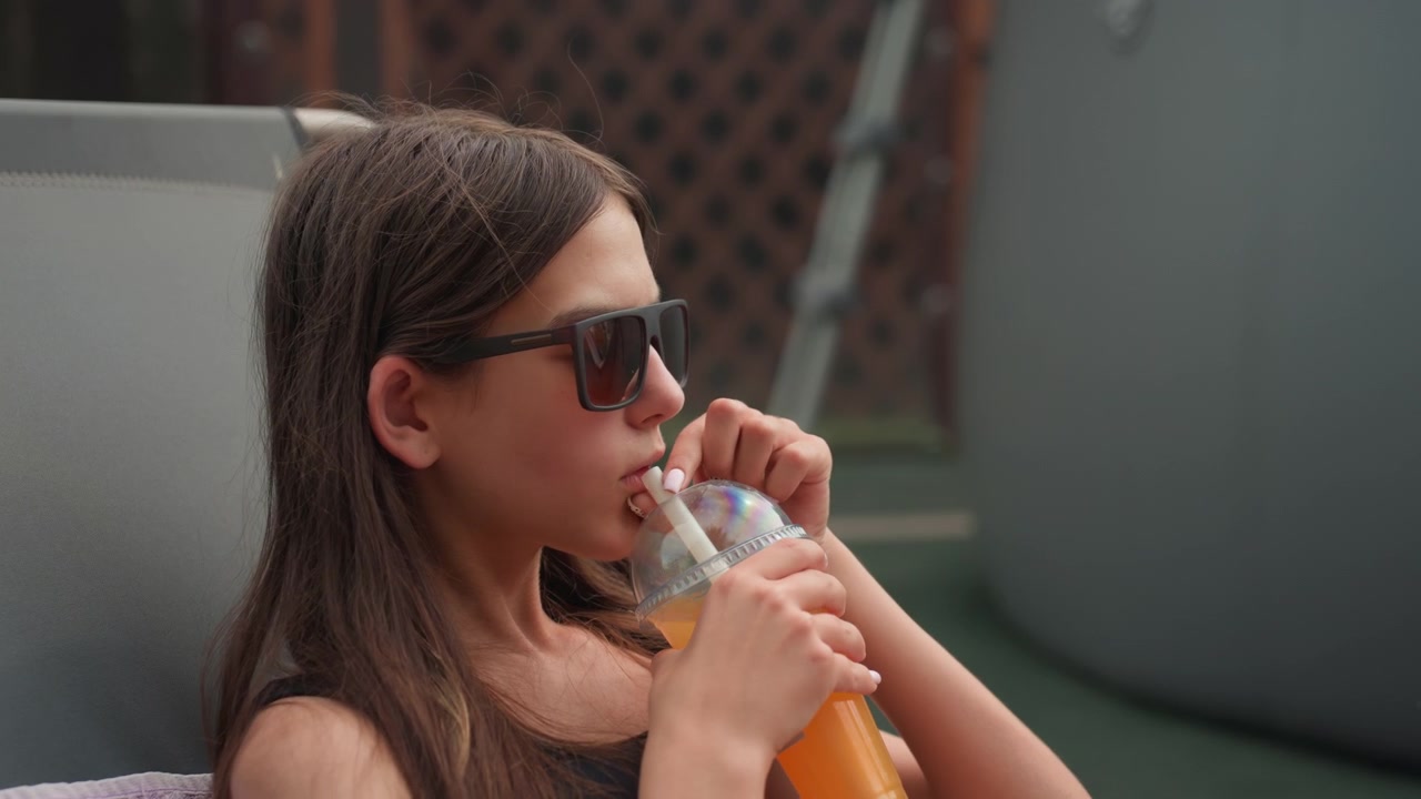 Young woman relaxes on gray outdoor chair reaching for and drinking orange juice from clear plastic cup with dome lid. Woman wears black sunglasses in urban backyard setting with greenery