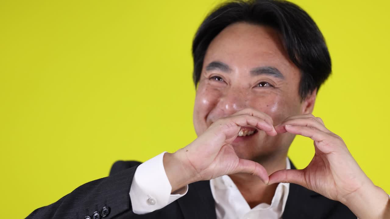 Smiling businessman in suit forms heart shape with hands, bright yellow background, studio lighting