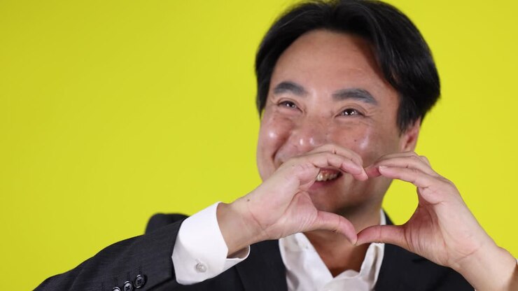 Uomo d'affari sorridente in giacca e cravatta che forma un cuore con le mani, sfondo giallo brillante, illuminazione da studio